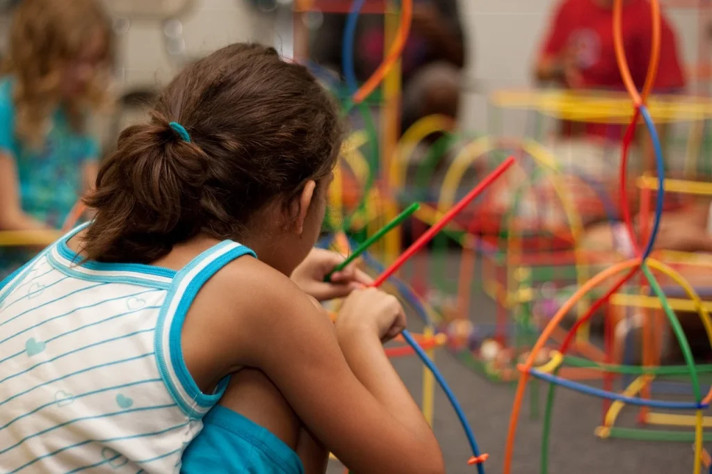 Image of a young female child playing with a toy facing away from the camera. Background includes a blur overlay showing more toys and children in the room in front of the child.