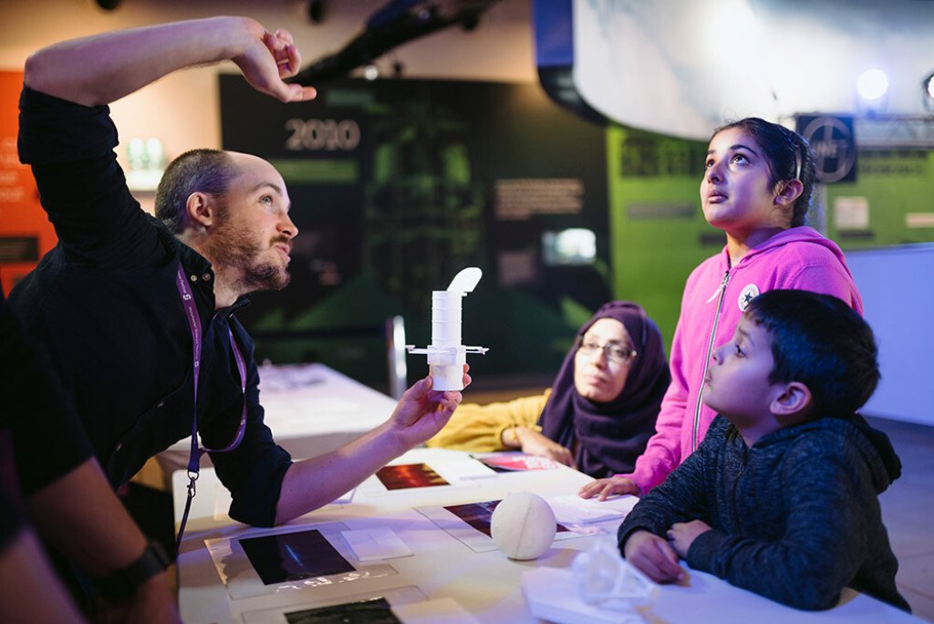 An image of four individuals, on adult man kneeling down, one adult woman kneeling down and two young children standing around a table watching the man describe something while holding an item.