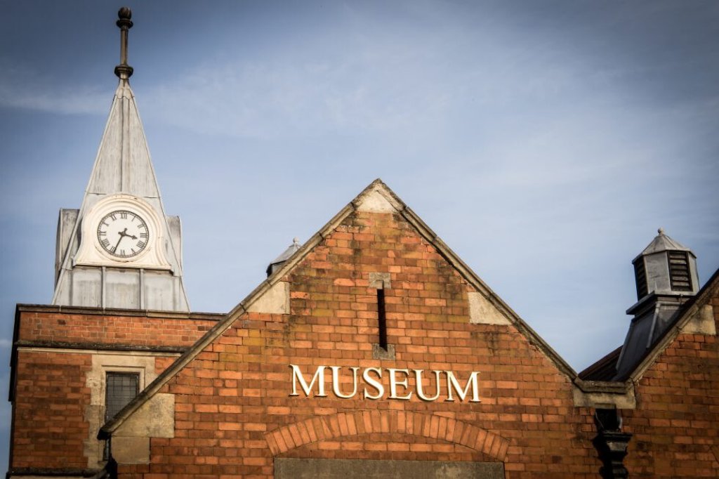 Image of a red brick museum building with the museum sign in front with blue skies behind it.