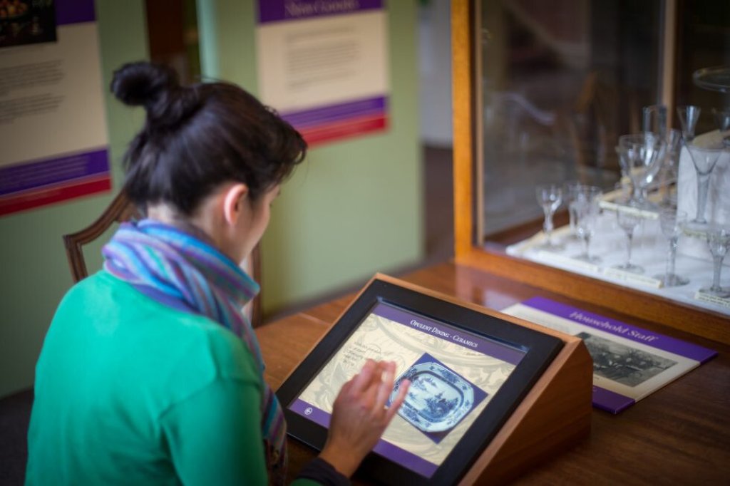 Woman in a green top using an interactive screen by a display case of drinking glasses.
