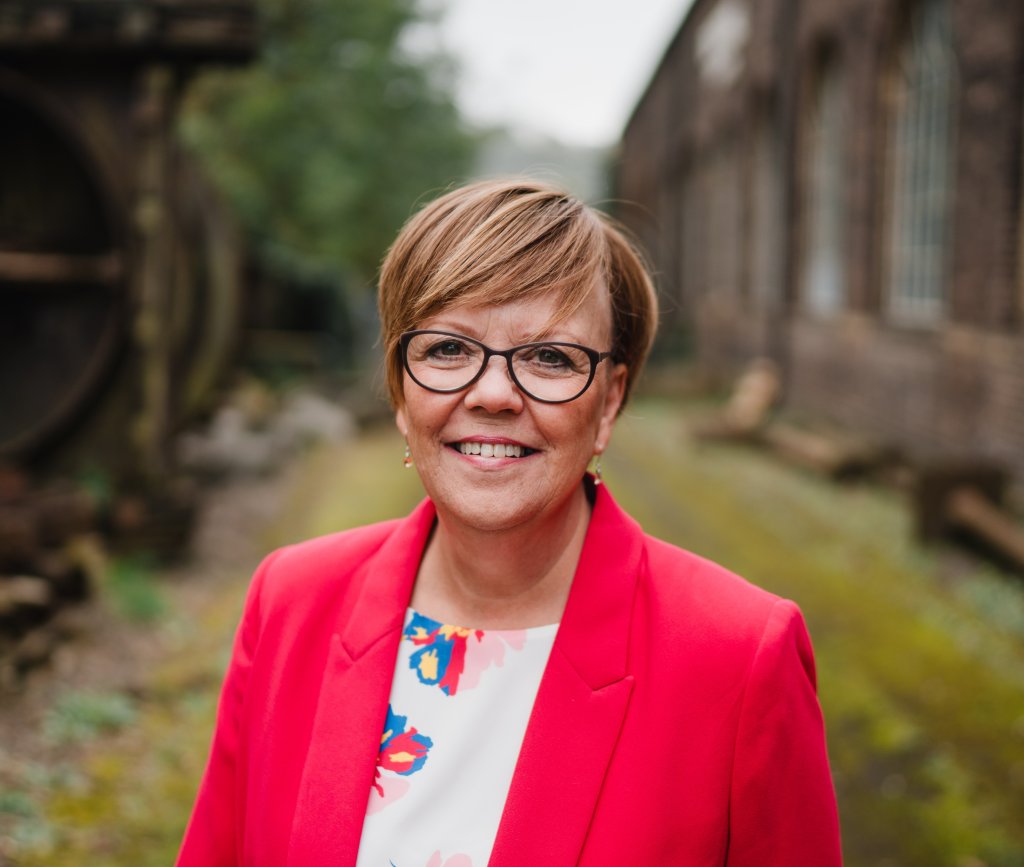 Karen wears glasses and has short light brown hair. She's wearing a pink jacket over a white patterned dress.