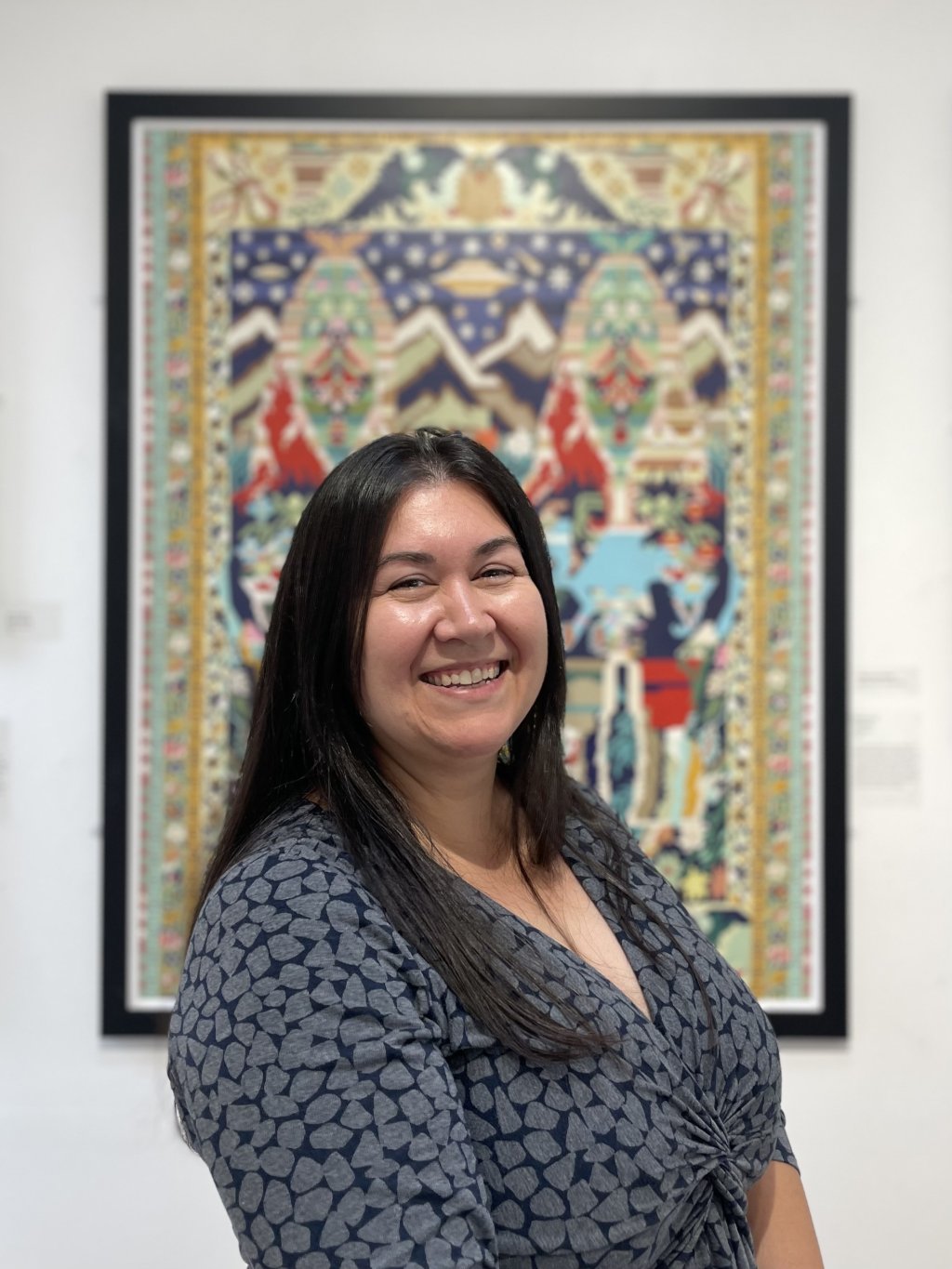Smiling woman with long dark hair in a blue and grey patterned top in front of a colourful picture.